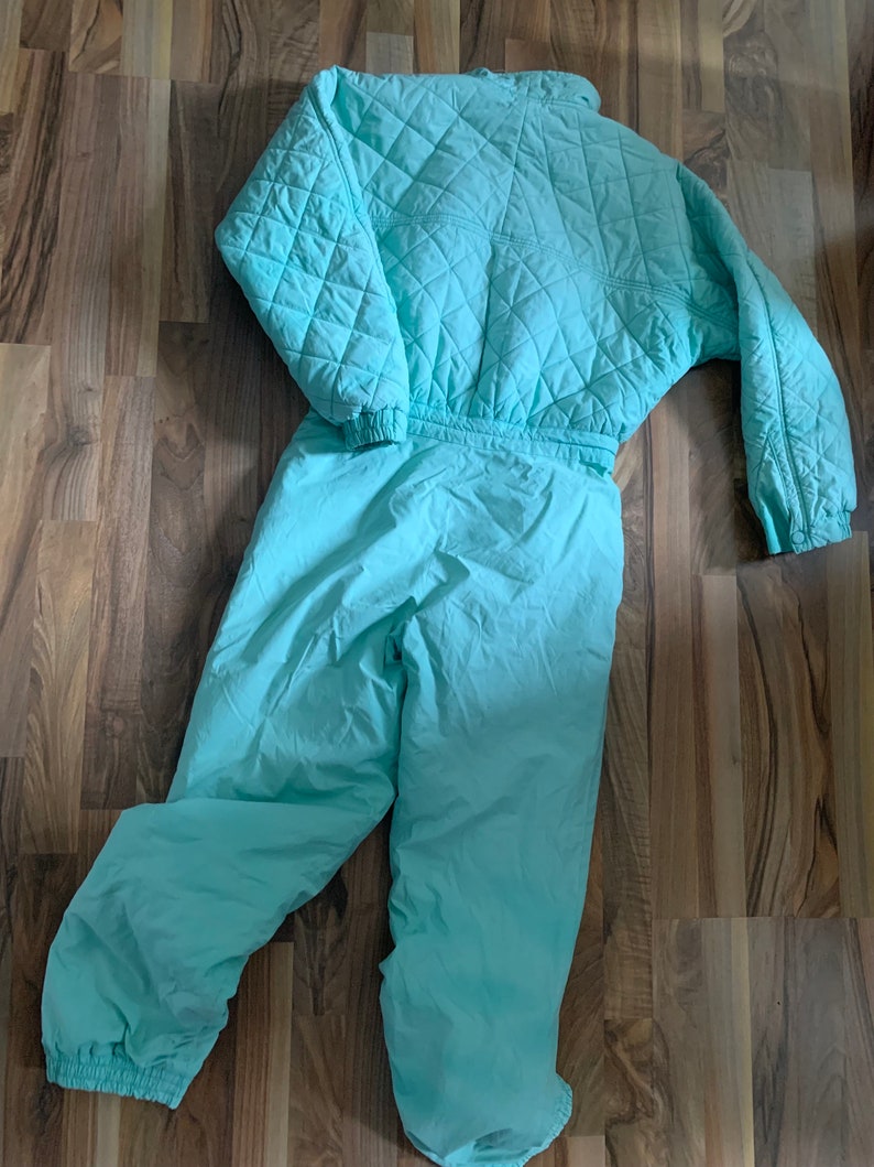 May include: A light blue quilted ski suit with a zipper closure. The suit has a quilted top and matching pants.