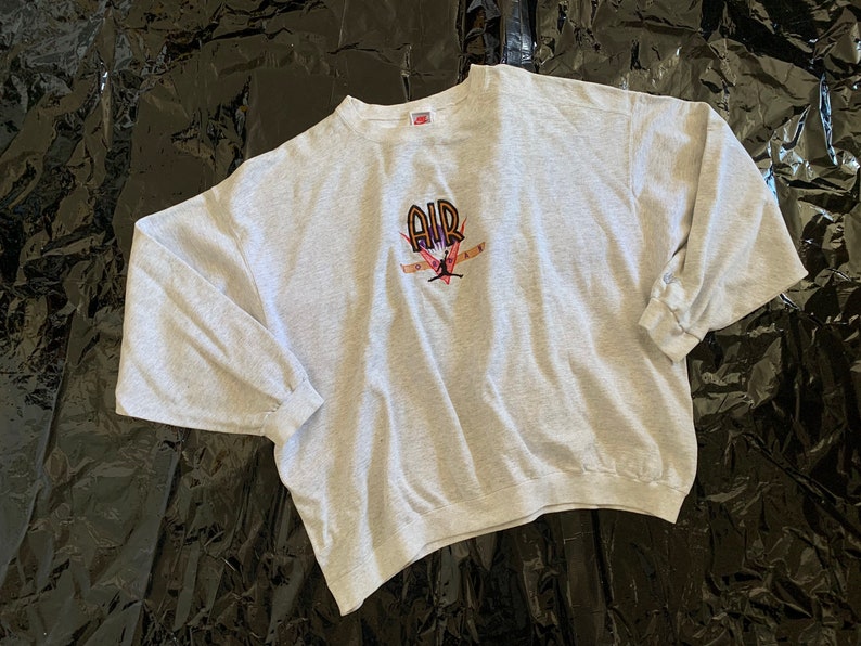 K&ouml;nnte beinhalten: Ein grauer Langarm-Sweatshirt mit einem Aufdruck eines Basketballspielers, der einen Dunk macht, mit dem Wort "AIR" &uuml;ber dem Spieler. Der Aufdruck ist in Schwarz, Orange und Pink.