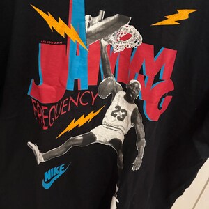 May include: Black t-shirt with a graphic of a basketball player mid-dunk. The design incorporates the words "JAM" and "FREQUENCY" in red and blue, with a Nike logo. The player is wearing a jersey with the number 23.