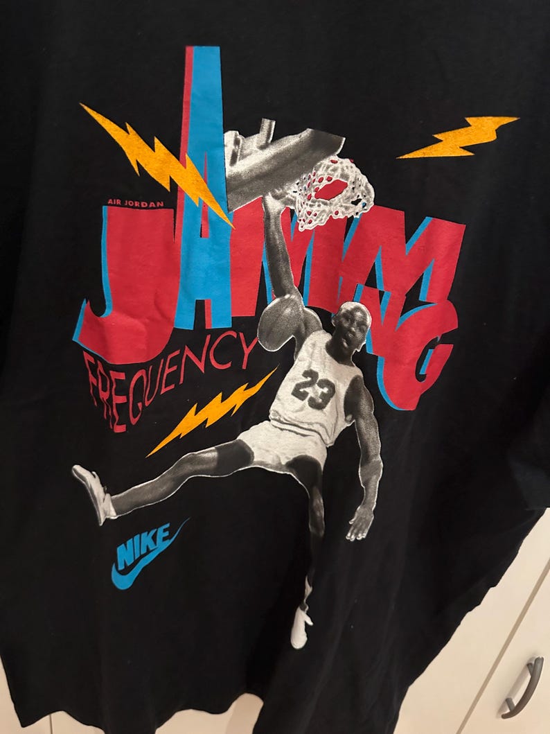 May include: Black t-shirt with a graphic of a basketball player mid-dunk. The design incorporates the words "JAM" and "FREQUENCY" in red and blue, with a Nike logo. The player is wearing a jersey with the number 23.