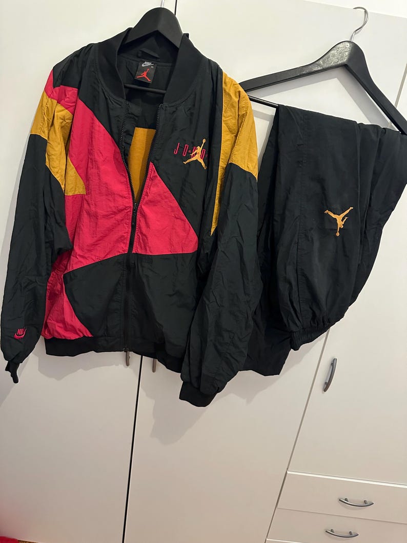 K&ouml;nnte beinhalten: Schwarzer, goldener und roter Nylon-Trainingsanzug. Die Jacke hat einen Rei&szlig;verschluss vorne und das Jordan-Logo. Die Hose ist schwarz mit einem goldenen Jordan-Logo. Die Jacke hat einen schwarzen Kragen und B&uuml;ndchen. Der Trainingsanzug h&auml;ngt an einem schwarzen Kleiderb&uuml;gel.