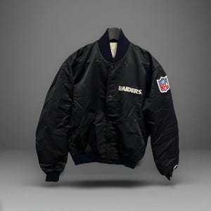 Könnte beinhalten: Schwarze Bomberjacke aus Satin mit dem Wort "Raiders" in weißer Schrift auf der Brust. Die Jacke hat einen marineblauen Kragen und Bündchen sowie einen NFL-Patch auf dem rechten Ärmel. Die Jacke ist zugeknöpft und hat zwei Vordertaschen.