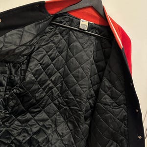 May include: A red, black, and white varsity jacket with a quilted black lining. The jacket is hanging on a black hanger. The collar and cuffs are red, and the body is black with white stripes.