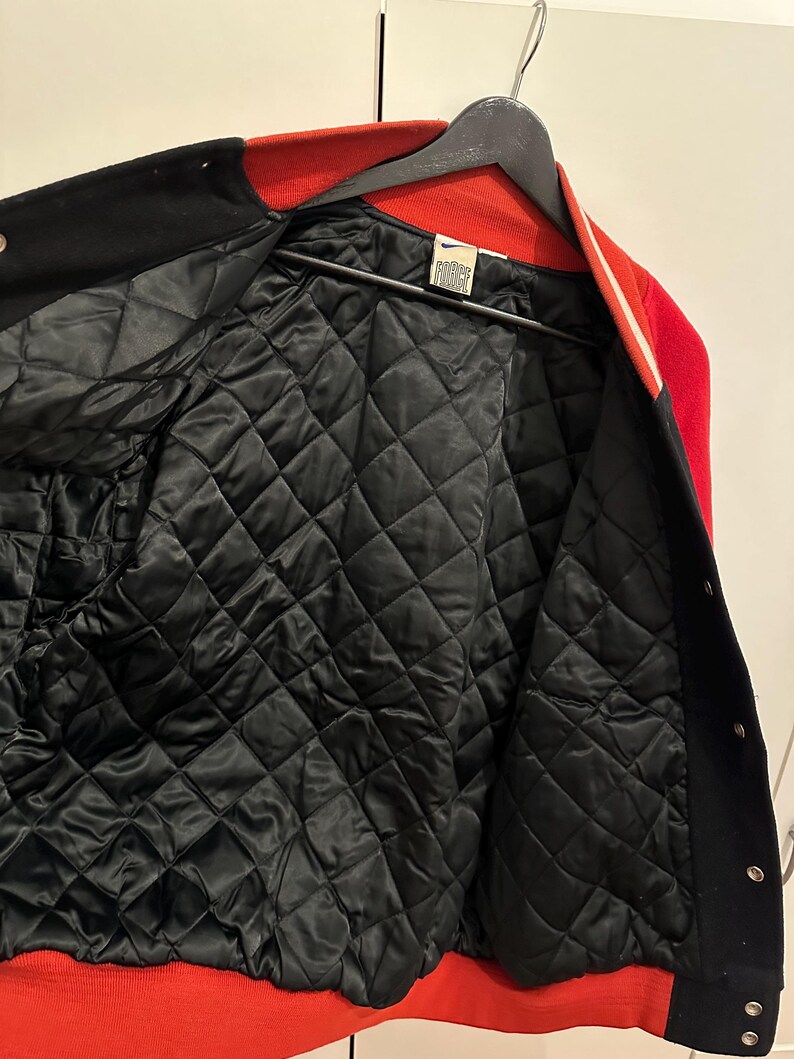 May include: A red, black, and white varsity jacket with a quilted black lining. The jacket is hanging on a black hanger. The collar and cuffs are red, and the body is black with white stripes.