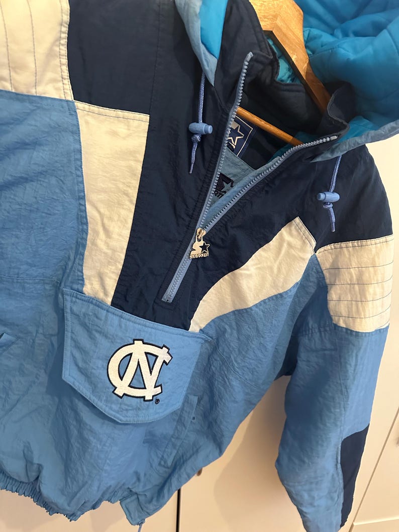 May include: A vintage Starter jacket in light blue, navy, and white. The jacket features a hood, a front pocket with a logo, and a zipper closure. The Starter logo is visible on the zipper pull.