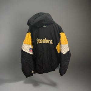 Könnte beinhalten: Schwarze Kapuzenjacke mit gelben und weißen Akzenten. Der Schriftzug "Steelers" in Gold und ein Teamlogo sind auf der Jacke zu sehen. Die Schultern sind gesteppt. Ein klassisches Sport-Thema Kleidungsstück.