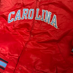 May include: Red satin bomber jacket with blue and black striped cuffs and collar. The jacket has a large blue and white "CAROLINA" logo on the front and a small "STARTER" logo on the upper left chest.
