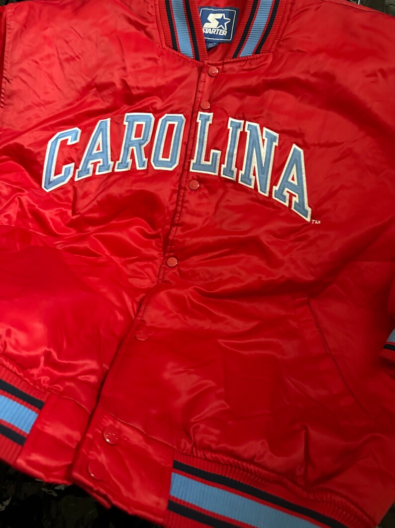 May include: Red satin bomber jacket with blue and black striped cuffs and collar. The jacket has a large blue and white "CAROLINA" logo on the front and a small "STARTER" logo on the upper left chest.