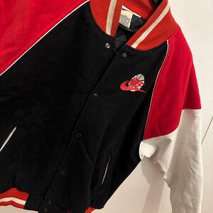 May include: A black, red, and white varsity jacket with a Nike logo featuring a basketball. The jacket has a red collar and sleeves, with white accents and a black body. The jacket is buttoned up and hanging on a black hanger.