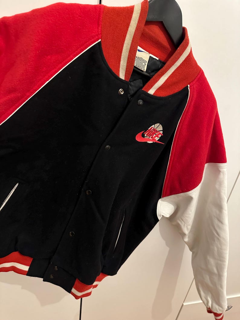 May include: A black, red, and white varsity jacket with a Nike logo featuring a basketball. The jacket has a red collar and sleeves, with white accents and a black body. The jacket is buttoned up and hanging on a black hanger.