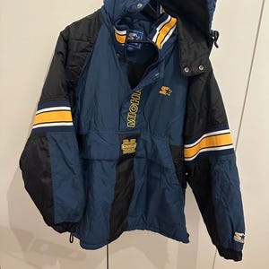 Könnte beinhalten: Blaue, schwarze und gelbe Starter-Jacke mit Kapuze. Die Jacke trägt den Schriftzug "MICHIGAN" und ein Logo auf der Vorderseite. Gelbe und weiße Streifen akzentuieren die Ärmel. Das Starter-Logo ist auf dem Ärmel sichtbar.