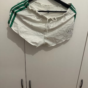 May include: White athletic shorts with green stripes on the sides. The shorts have a drawstring waist and a small logo on the leg. The shorts are hanging on a black hanger.