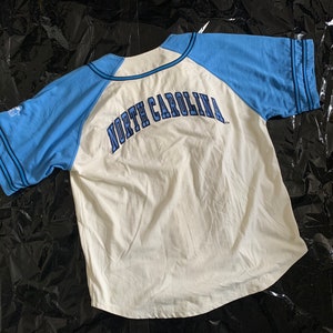 May include: White baseball jersey with blue sleeves and the text "NORTH CAROLINA" in blue on the back.