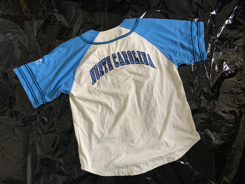 May include: White baseball jersey with blue sleeves and the text "NORTH CAROLINA" in blue on the back.