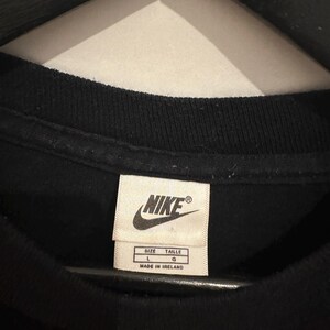 May include: Close-up of a black t-shirt collar with a Nike label. The label is white with black text, including the Nike logo, size information (L, G), and "MADE IN IRELAND".