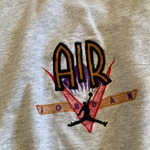 K&ouml;nnte beinhalten: Grauer Sweatshirt mit einem gestickten Logo, das das Wort "AIR" in orangefarbenen und schwarzen Buchstaben mit einem violetten Umriss zeigt. Das Logo enth&auml;lt auch einen Basketballspieler in schwarzer Silhouette, der mit einem Basketball springt. Der Text "JORDAN" ist in orangefarbenen und schwarzen Buchstaben unter dem Logo gestickt.