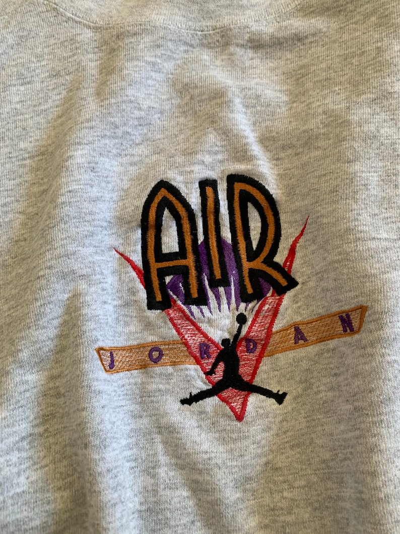 K&ouml;nnte beinhalten: Grauer Sweatshirt mit einem gestickten Logo, das das Wort "AIR" in orangefarbenen und schwarzen Buchstaben mit einem violetten Umriss zeigt. Das Logo enth&auml;lt auch einen Basketballspieler in schwarzer Silhouette, der mit einem Basketball springt. Der Text "JORDAN" ist in orangefarbenen und schwarzen Buchstaben unter dem Logo gestickt.