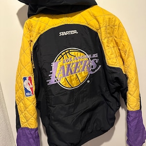 Puede incluir: Una chaqueta negra y amarilla de la marca Starter con un logotipo de los Los Angeles Lakers en morado y amarillo en la espalda. La chaqueta tiene capucha y un diseño acolchado.
