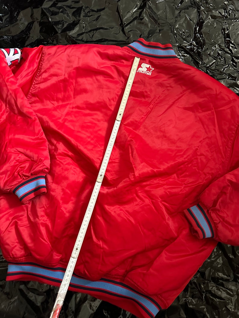May include: Red satin baseball jacket with blue and black trim. The jacket has a large "STARTER" logo on the back.