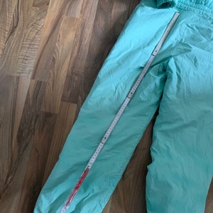 May include: A pair of light blue ski pants with elastic cuffs. The pants are laid flat on a wooden floor with a measuring tape.