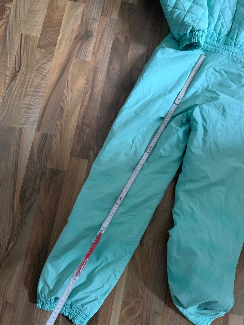 May include: A pair of light blue ski pants with elastic cuffs. The pants are laid flat on a wooden floor with a measuring tape.