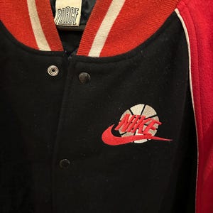 May include: Black and red varsity jacket with the word "FORCE" on the collar. The jacket features a red and white striped collar and cuffs. The Nike logo, a red swoosh with a basketball design, is embroidered on the front.