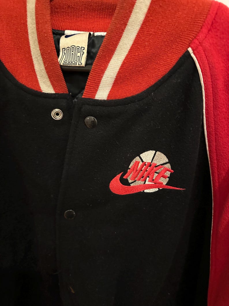 May include: Black and red varsity jacket with the word "FORCE" on the collar. The jacket features a red and white striped collar and cuffs. The Nike logo, a red swoosh with a basketball design, is embroidered on the front.