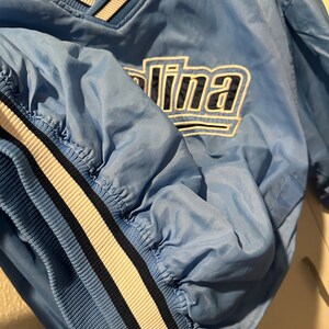 K&ouml;nnte beinhalten: Eine hellblaue Jacke der Marke Starter mit wei&szlig;en und dunkelblauen Streifen an Kragen und &Auml;rmeln. Die Jacke hat ein gro&szlig;es gesticktes Carolina-Logo auf der Vorderseite.