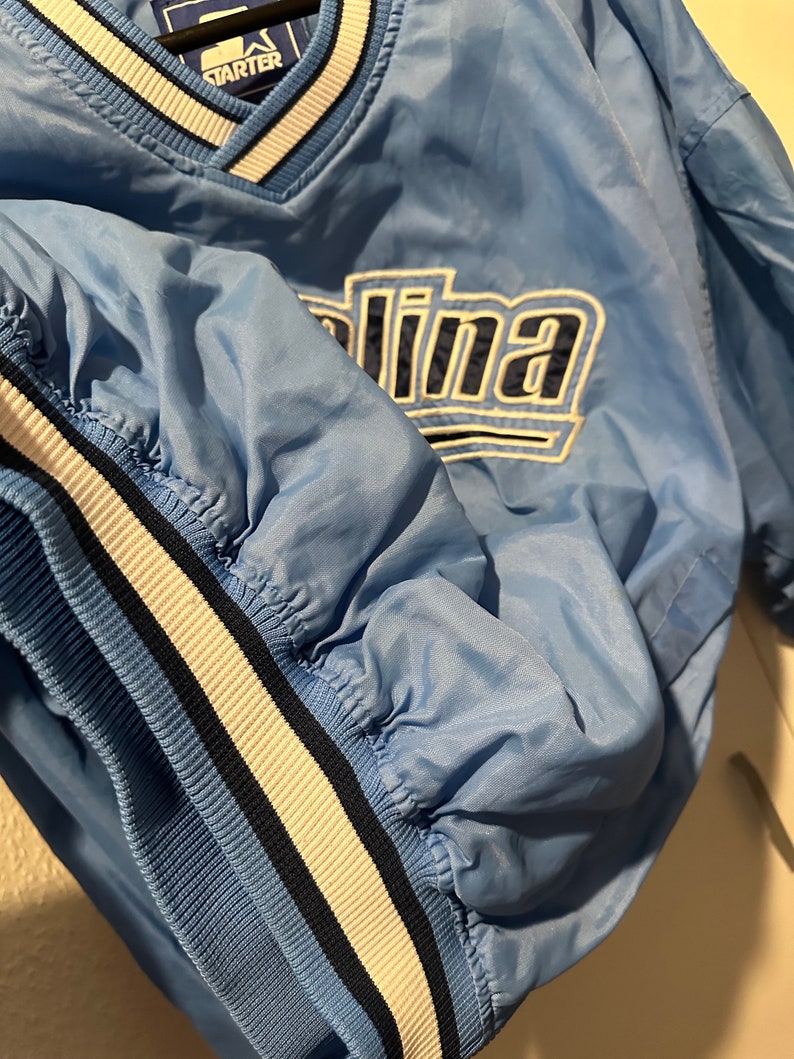 K&ouml;nnte beinhalten: Eine hellblaue Jacke der Marke Starter mit wei&szlig;en und dunkelblauen Streifen an Kragen und &Auml;rmeln. Die Jacke hat ein gro&szlig;es gesticktes Carolina-Logo auf der Vorderseite.