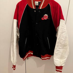 May include: Black, red, and white varsity jacket with a basketball logo. The jacket has a black body, red sleeves, and white sleeves. The collar and cuffs are red and white striped. The Nike logo is on the chest.