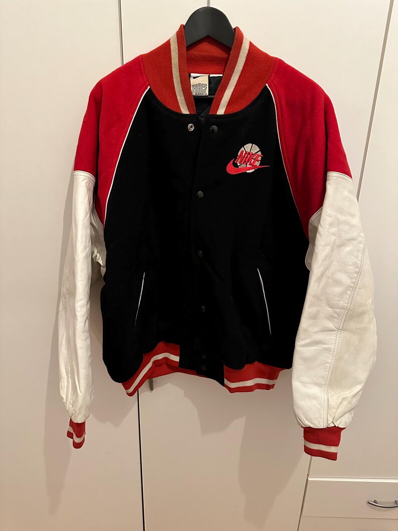 May include: Black, red, and white varsity jacket with a basketball logo. The jacket has a black body, red sleeves, and white sleeves. The collar and cuffs are red and white striped. The Nike logo is on the chest.