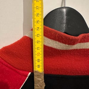 May include: A red, black, and white athletic jacket is hanging on a black hanger. A yellow measuring tape is taped to the jacket, showing a measurement of approximately 27 inches.