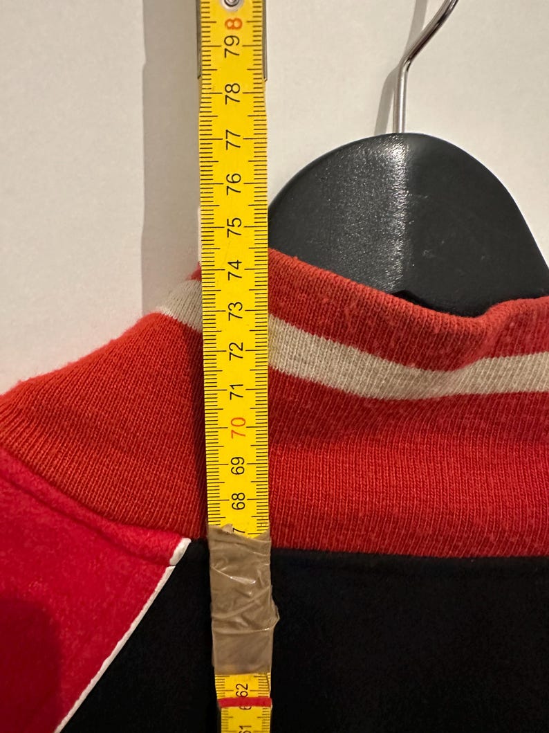 May include: A red, black, and white athletic jacket is hanging on a black hanger. A yellow measuring tape is taped to the jacket, showing a measurement of approximately 27 inches.