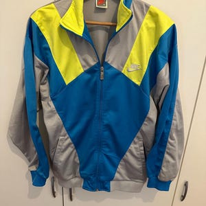 Könnte beinhalten: Vintage Nike Trainingsjacke in Grau, Blau und Gelb. Die Jacke hat einen durchgehenden Reißverschluss, einen Stehkragen und das Nike-Logo auf der Brust. Das Design umfasst farbblockierte Einsätze und gerippte Bündchen und Saum.
