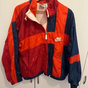Könnte beinhalten: Eine Vintage Nike Windjacke im Colorblock-Design. Die Jacke hat Einsätze in Rot, Orange und Marineblau. Sie hat einen durchgehenden Reißverschluss, ein weißes Nike-Logo und eine Kapuze. Die Jacke hängt an einem Holzkleiderbügel.