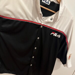 Peut inclure: Une chemise boutonn&eacute;e Fila blanche, noire et rouge. La chemise a une section sup&eacute;rieure en mesh et une section inf&eacute;rieure noire avec le logo Fila sur la poitrine. La chemise est suspendue &agrave; un cintre en bois.