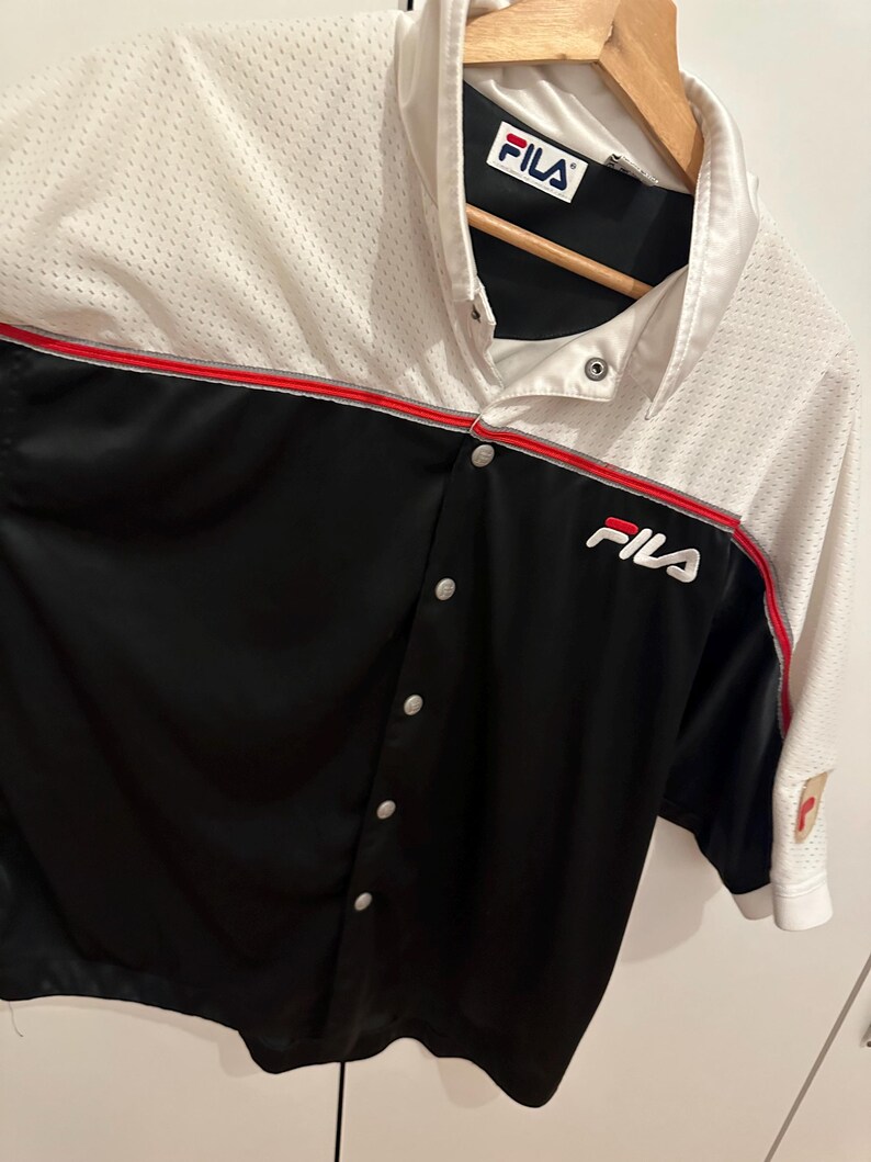 Peut inclure: Une chemise boutonn&eacute;e Fila blanche, noire et rouge. La chemise a une section sup&eacute;rieure en mesh et une section inf&eacute;rieure noire avec le logo Fila sur la poitrine. La chemise est suspendue &agrave; un cintre en bois.