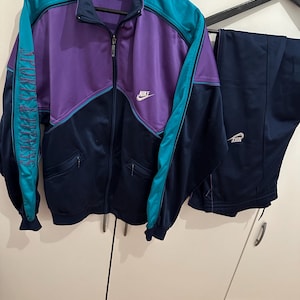 May include: A vintage Nike tracksuit in a color block design. The jacket is purple, teal, and navy blue with the Nike logo on the chest. The matching pants are navy blue with the Nike logo on the left leg. The jacket has the text "SPORTSWEAR" on the left sleeve.