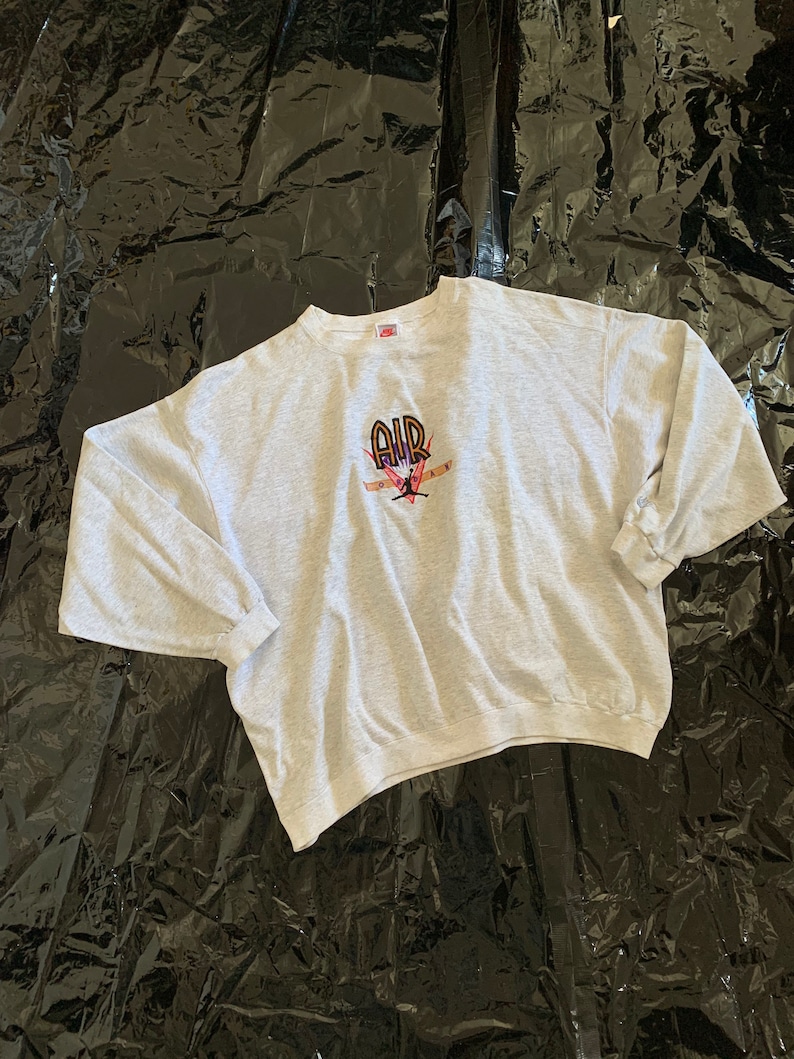 K&ouml;nnte beinhalten: Ein grauer Pullover mit einem schwarzen, orangefarbenen und roten gestickten Motiv eines Basketballspielers mit dem Wort "AIR" &uuml;ber dem Spieler.