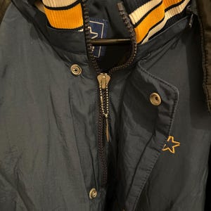 May include: Navy blue hooded jacket with a gold star emblem. The jacket features a full zipper, snap closures, and a striped collar in navy, gold, and white. The jacket is made of a shiny, water-resistant material.