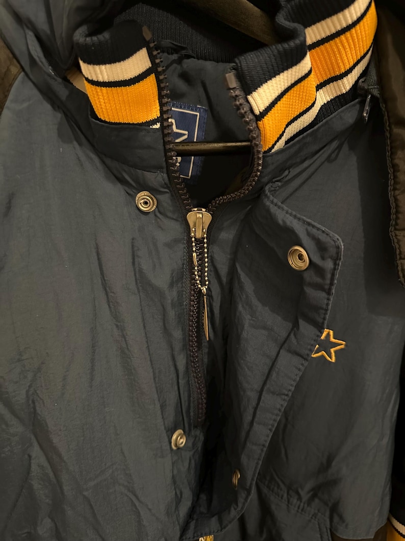 May include: Navy blue hooded jacket with a gold star emblem. The jacket features a full zipper, snap closures, and a striped collar in navy, gold, and white. The jacket is made of a shiny, water-resistant material.