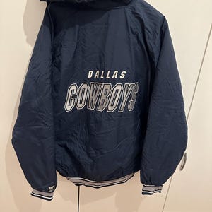 Könnte beinhalten: Marineblaue Kapuzenjacke mit dem Text "DALLAS COWBOYS" in weißen und silbernen Buchstaben auf dem Rücken. Die Jacke hat einen weiß-grau gestreiften Besatz an den Bündchen und am Saum.