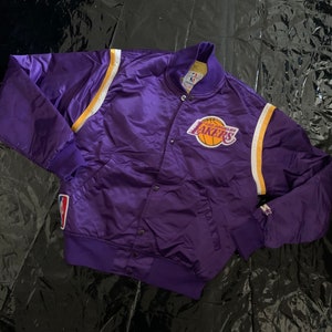 Könnte beinhalten: Eine violette Satin-Bomberjacke mit gelben und weißen Streifen an den Ärmeln. Die Jacke hat ein großes Los Angeles Lakers Logo auf der Vorderseite. Die Jacke ist in gutem Zustand und hat einen Vintage-Look.