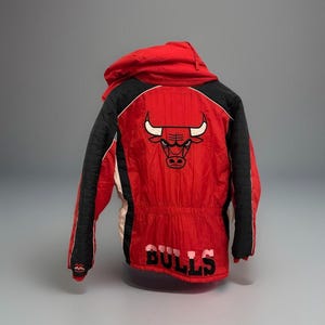 Peut inclure: Veste rouge et noire des Chicago Bulls avec capuche. La veste présente le logo des Bulls au dos et le mot "BULLS" en lettres blanches. La veste a une bordure noire et blanche.
