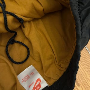 K&ouml;nnte beinhalten: Schwarze Nike-Sporthose mit senfgelbem Innenfutter. Die Hose hat einen Kordelzug und ein Etikett mit dem Nike-Logo und Produktinformationen. Hergestellt aus Baumwolle, Polyester und Nylon.