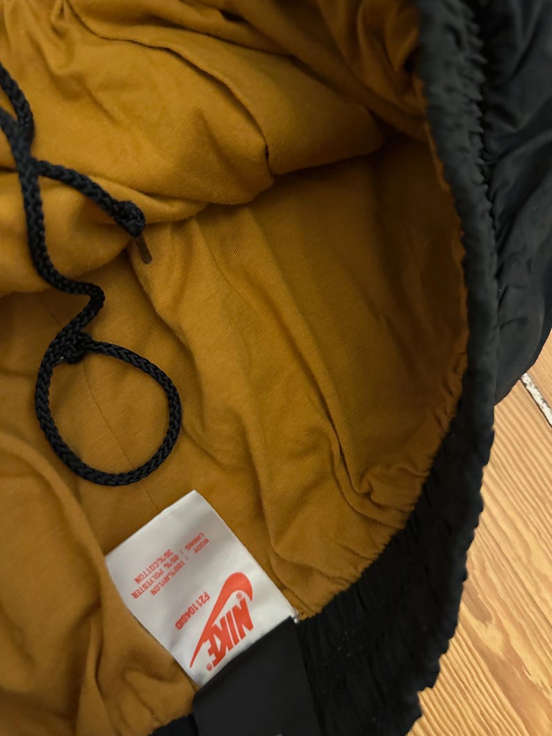 K&ouml;nnte beinhalten: Schwarze Nike-Sporthose mit senfgelbem Innenfutter. Die Hose hat einen Kordelzug und ein Etikett mit dem Nike-Logo und Produktinformationen. Hergestellt aus Baumwolle, Polyester und Nylon.