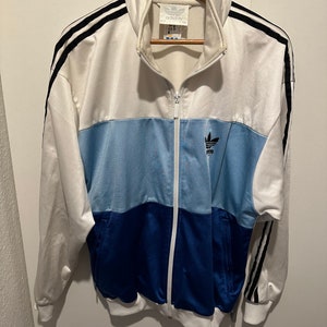 Op de afbeelding: Een wit, blauw en donkerblauw Adidas trainingsjack met zwarte strepen op de mouwen en een klein Adidas logo op de borst.