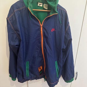 Könnte beinhalten: Eine marineblaue Nike Kapuzenjacke mit orangefarbenem Reißverschluss und Nähten. Die Jacke hat eine grüne Kapuze und grüne Akzente an den Taschen und Manschetten. Ein rotes Nike-Logo befindet sich auf der Brust. Die Jacke hängt an einem Holzbügel.