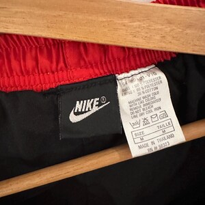 May include: Black Nike athletic shorts with a red waistband. The tag says "SHELL: 100% POLYESTER, LINING: 65% POLYESTER, 35% COTTON, MACHINE WASH COLD WITH LIKE COLORS, REMOVE IMMEDIATELY, DO NOT BLEACH, LINE DRY COOL IRON, SIZE M, TAILLE M, MADE IN THAILAND, RN #56323".