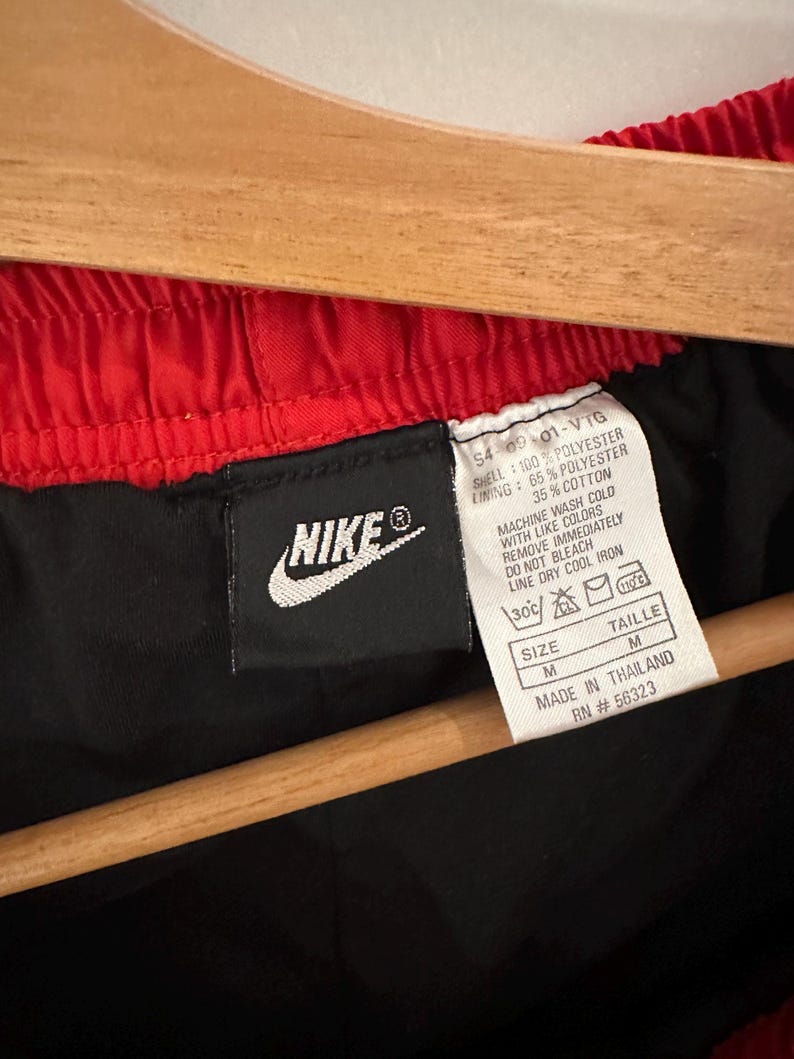 May include: Black Nike athletic shorts with a red waistband. The tag says "SHELL: 100% POLYESTER, LINING: 65% POLYESTER, 35% COTTON, MACHINE WASH COLD WITH LIKE COLORS, REMOVE IMMEDIATELY, DO NOT BLEACH, LINE DRY COOL IRON, SIZE M, TAILLE M, MADE IN THAILAND, RN #56323".